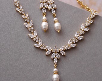 May include: A gold-coloured jewellery set comprising a necklace and matching earrings. The necklace and earrings feature a leaf-like design with clear crystals and pear-shaped white pearls. The set is displayed on a grey surface.