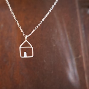 May include: A silver-colored necklace featuring a small house pendant. The pendant is a minimalist design, with a house outline and a rectangular door. The necklace hangs against a brown background.
