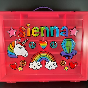 May include: Pink plastic storage box with a purple handle. The box is decorated with colorful drawings of a rainbow, a unicorn, a diamond, stars, peace signs, and hearts. The name "sienna" is written in rainbow letters across the top.