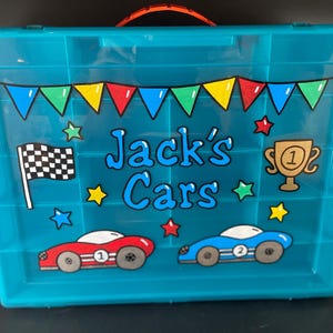 May include: A turquoise plastic storage bin with a handle and a decorated front. The front features the text "Jack's Cars" in blue, along with illustrations of race cars, a checkered flag, and a trophy. Colorful star and banner accents complete the design.
