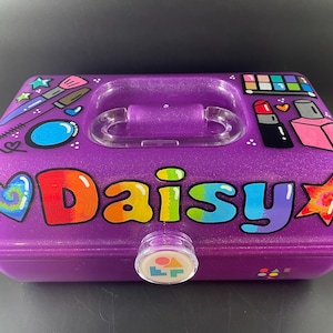 May include: Purple makeup case with a clear handle and clasp. The case is decorated with colorful illustrations of makeup items and the name "Daisy" in rainbow letters. A heart and star are also painted on the case.