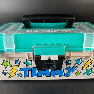 May include: A teal and off-white plastic toolbox with a black handle and clear lid. The toolbox is decorated with stars, lightning bolts, and the name "TIMMY" in blue letters. The brand name "SHEFFIELD" is visible on the lid.