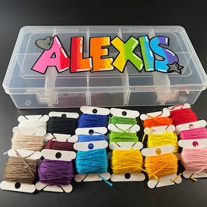 May include: A clear plastic organizer box with multiple compartments, labeled "ALEXIS" in colorful graffiti-style lettering. Below, spools of embroidery floss in various colors, including white, black, blue, green, yellow, orange, red, and pink.
