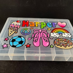 May include: A clear plastic storage box with 24 compartments. The lid is decorated with colorful drawings of an ice cream cone, a soccer ball, a peace sign, a rainbow, a donut, ballet shoes, and stars. The name "Harper" is written in colorful letters across the top.