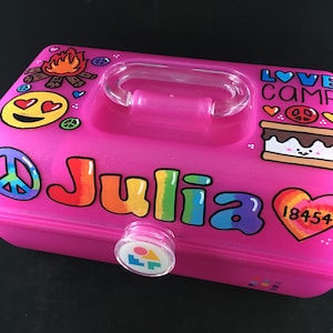 May include: Pink plastic storage box with a clear handle and a colorful design. The box has a peace sign, a smiley face, a campfire, a s'more, a heart with the text "18454", and the text "LOVE CAMP" and "Julia".