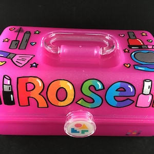 May include: Pink plastic makeup case with a clear handle and a rainbow glitter design. The case has a clear lid with a pink button and the word "Rose" written in rainbow letters.