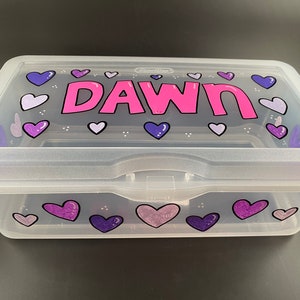 May include: A clear plastic storage box with a pink and purple heart design. The name "DAWN" is written in pink letters on the lid.
