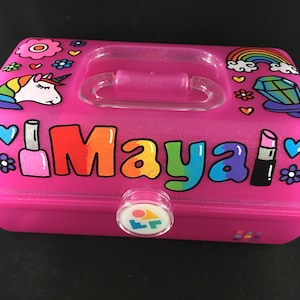 May include: Pink plastic storage box with a clear lid and a handle. The box has a rainbow glitter finish and is decorated with a unicorn, a rainbow, a diamond, lipstick, nail polish, and flowers. The name "Maya" is written in rainbow letters on the front of the box.