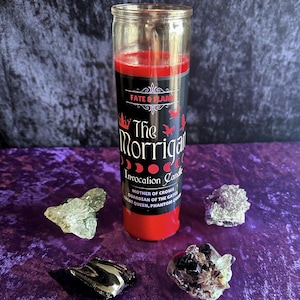 May include: A red candle with a black label that reads "The Morrigan Invocation Candle" with the text "Fate & Flame" at the top. The label also includes the text "Mother of Crows, Guardian of the Gates, Great Queen, Phantom Queen".