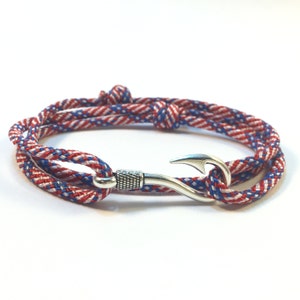 May include: A nautical-themed bracelet featuring a red, white, and blue braided rope design. The bracelet is secured with a silver-colored hook clasp. The rope is knotted in several places, adding to the bracelet's unique style.