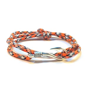 May include: A bracelet featuring a woven rope design in orange, gray, and black. The bracelet has a silver-colored hook clasp. The rope is wrapped around the wrist twice.