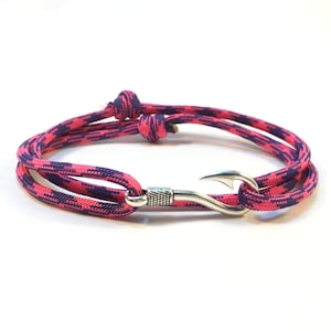 May include: A pink and purple rope bracelet with a silver-colored hook clasp. The bracelet is made of woven nylon rope and features a repeating pattern of pink and purple. The hook clasp is detailed with a textured design.