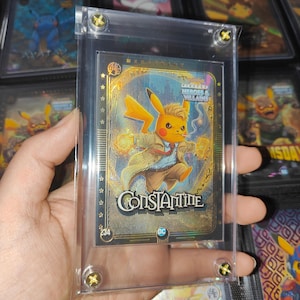 May include: A collectible trading card encased in a clear magnetic acrylic holder. The card features a yellow Pikachu character dressed as John Constantine, wearing a tan trench coat, blue shirt, and red tie, with the text Constantine and DC logo.