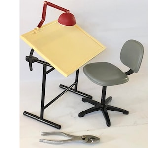 May include: A miniature drafting table set with a yellow tabletop, red lamp, and grey chair. The table has black legs and a tilting surface. A pair of pliers is in the foreground. The set is designed for a dollhouse or model.