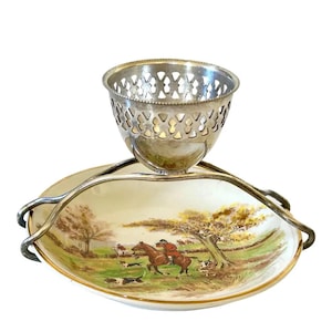 May include: An antique silver-plated egg cup and serving dish. The dish features a pastoral scene with a rider on horseback, dogs, and trees. The egg cup has a decorative pierced design.