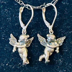May include: Silver-toned angel earrings with detailed wings and lever-back closures. The earrings feature small, detailed cherub figures. The earrings are suspended from a delicate silver chain against a dark background.