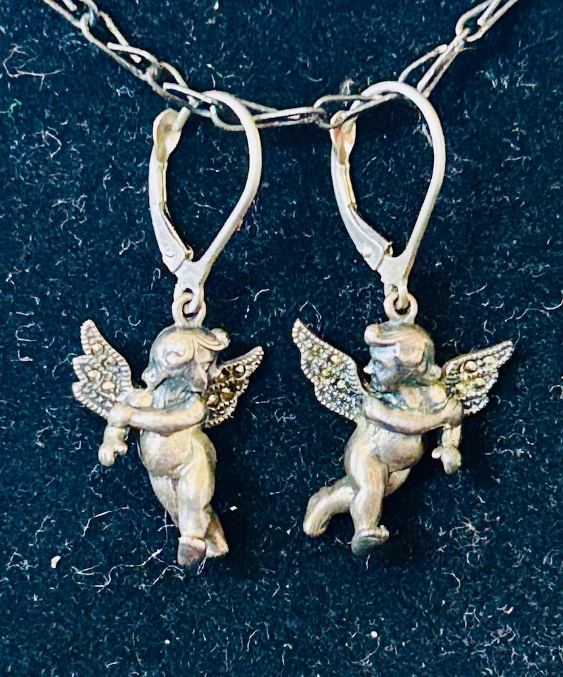 May include: Silver-toned angel earrings with detailed wings and lever-back closures. The earrings feature small, detailed cherub figures. The earrings are suspended from a delicate silver chain against a dark background.