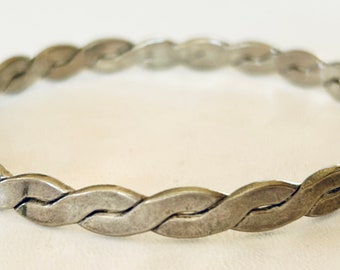 May include: A silver-toned bracelet with a braided design. The bracelet is made of interlocking, oval-shaped links that create a woven pattern. The jewelry is displayed on a white surface.