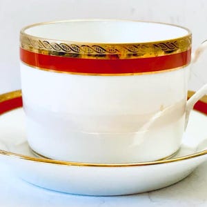 Może przedstawiać: Biały zestaw filiżanki i spodka ze złotymi zdobieniami i czerwonym paskiem. Filiżanka ma zakrzywiony uchwyt i stoi na pasującym spodku. Złote wykończenie ma powtarzający się wzór. Klasyczny design do herbaty lub kawy.
