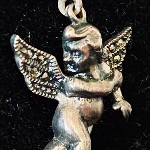 May include: A silver-toned earring featuring a cherubic angel with detailed wings. The angel is depicted in a standing pose, with its arms slightly outstretched. The earring has a hook closure.