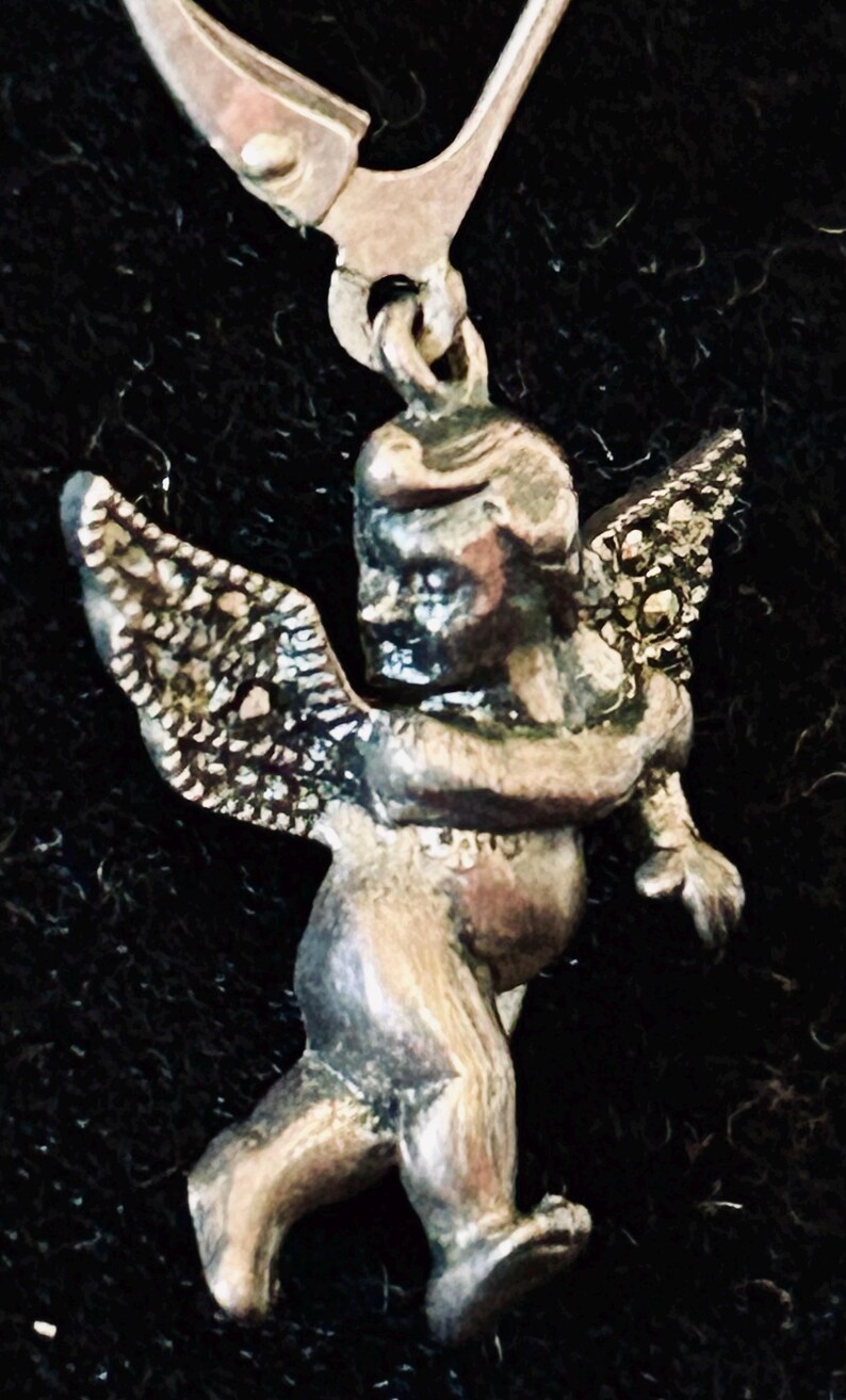 May include: A silver-toned earring featuring a cherubic angel with detailed wings. The angel is depicted in a standing pose, with its arms slightly outstretched. The earring has a hook closure.