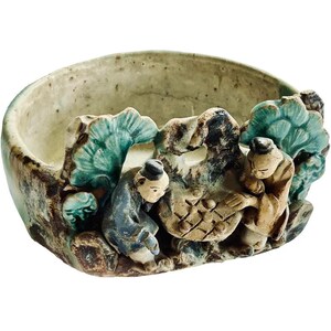 Puede incluir: Un cuenco de cerámica con un relieve detallado de figuras y follaje. El cuenco presenta un interior verde claro y un exterior texturizado con acentos azules, marrones y beiges. Las figuras visten trajes tradicionales.