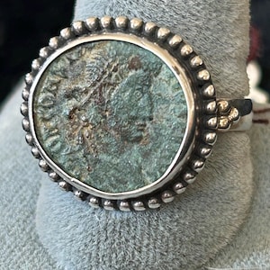 Op de afbeelding: Een zilveren ring met een antieke munt met een gedetailleerd profiel. De munt heeft een verweerde, groen getinte patina, gezet in een kralenrand. De ringband is eenvoudig en elegant, en benadrukt het ingewikkelde ontwerp van de munt.