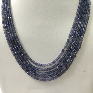 K&ouml;nnte beinhalten: Eine mehrstr&auml;ngige Halskette aus kleinen, facettierten, dunkelblauen Perlen. Die Halskette ist auf einer wei&szlig;en Mannequin-B&uuml;ste drapiert, die ihr mehrschichtiges Design und die subtilen Farbvariationen der Perlen hervorhebt.