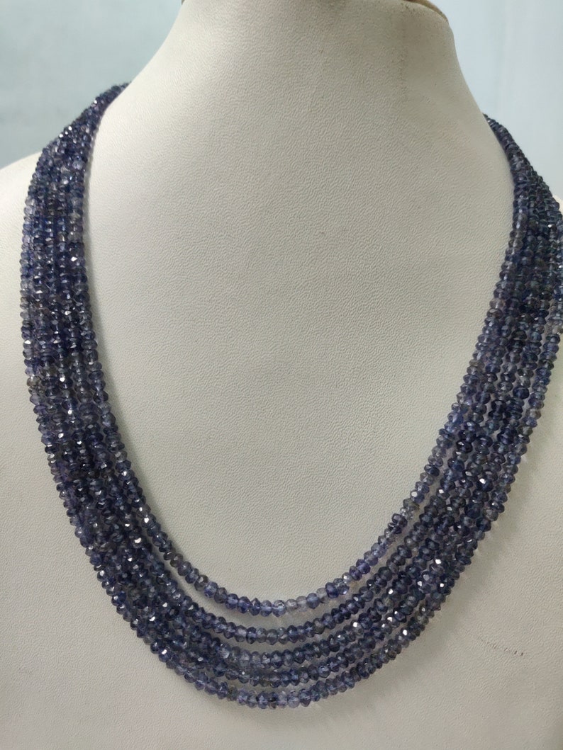 K&ouml;nnte beinhalten: Eine mehrstr&auml;ngige Halskette aus kleinen, facettierten, blau-violetten Perlen. Die Halskette ist auf einer wei&szlig;en Mannequin-B&uuml;ste drapiert, die ihr mehrschichtiges Design und ihre Farbe hervorhebt. Die Perlen haben einen subtilen Glanz.