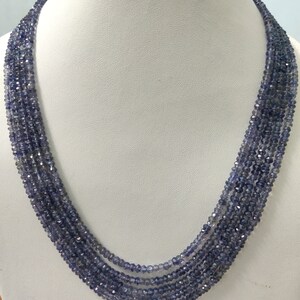 K&ouml;nnte beinhalten: Eine mehrstr&auml;ngige Halskette aus kleinen, facettierten, blau-violetten Perlen. Die Halskette ist auf einer wei&szlig;en Mannequin-B&uuml;ste drapiert, die ihr mehrschichtiges Design und ihre Farbe hervorhebt. Die Perlen haben einen subtilen Glanz.