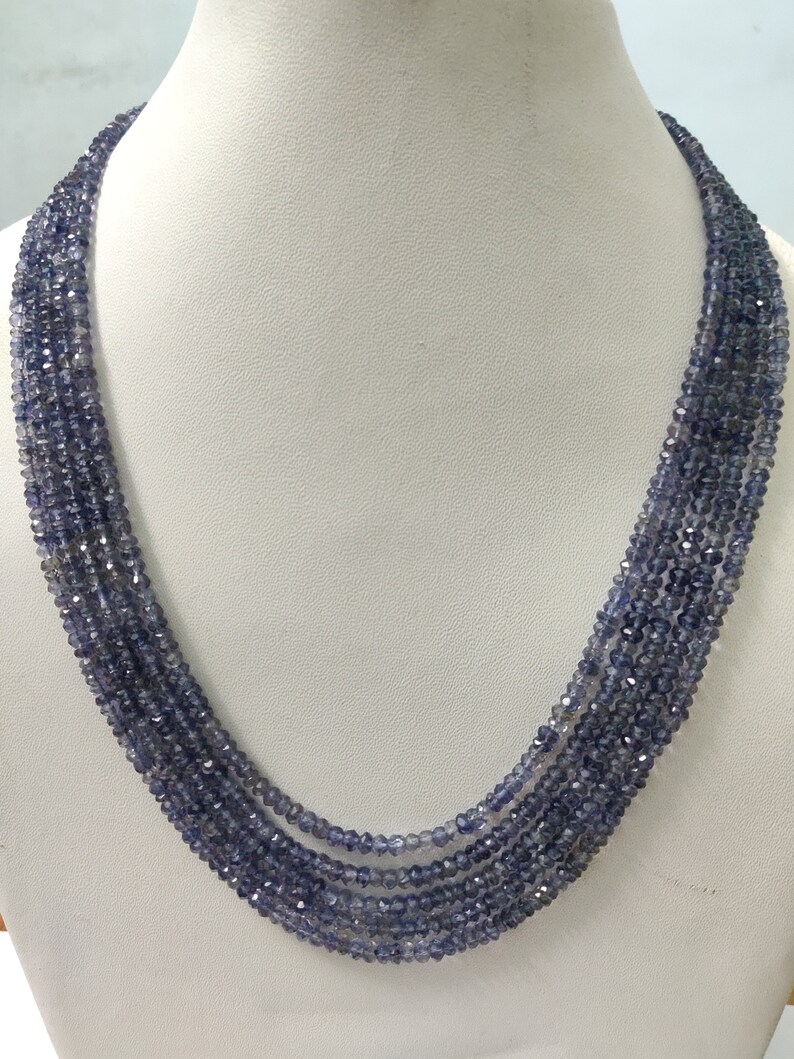 K&ouml;nnte beinhalten: Eine mehrstr&auml;ngige Halskette aus kleinen, facettierten, blau-violetten Perlen. Die Halskette ist auf einer wei&szlig;en Mannequin-B&uuml;ste drapiert, die ihr mehrschichtiges Design und ihre Farbe hervorhebt. Die Perlen haben einen subtilen Glanz.