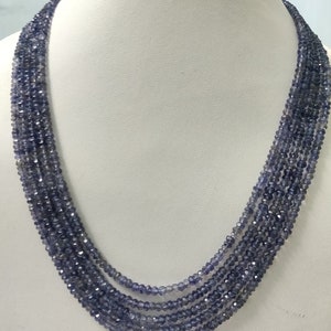 K&ouml;nnte beinhalten: Eine mehrstr&auml;ngige Halskette aus kleinen, facettierten, blau-violetten Perlen. Die Halskette wird auf einer wei&szlig;en Schaufensterpuppe pr&auml;sentiert, die ihr mehrschichtiges Design und ihre Farbe hervorhebt. Die Perlen fangen das Licht ein und erzeugen einen funkelnden Effekt.