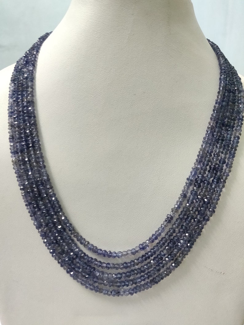 K&ouml;nnte beinhalten: Eine mehrstr&auml;ngige Halskette aus kleinen, facettierten, blau-violetten Perlen. Die Halskette wird auf einer wei&szlig;en Schaufensterpuppe pr&auml;sentiert, die ihr mehrschichtiges Design und ihre Farbe hervorhebt. Die Perlen fangen das Licht ein und erzeugen einen funkelnden Effekt.