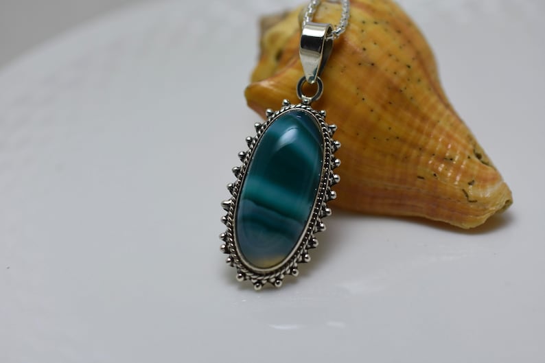 May include: A silver pendant with a green and white agate stone set in a silver oval frame with a decorative border. The pendant is hanging from a silver chain.