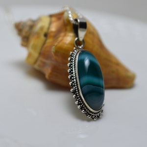 May include: A silver pendant with a green agate stone set in a silver bezel. The stone has white stripes and is oval shaped. The pendant is hanging from a silver chain.