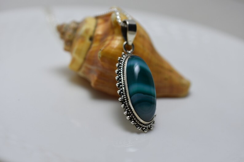 May include: A silver pendant with a green agate stone set in a silver bezel. The stone has white stripes and is oval shaped. The pendant is hanging from a silver chain.