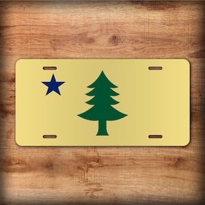 May include: A rectangular license plate with a gold background. The design features a dark blue star in the upper left corner and a green pine tree on the right. The plate has four mounting holes.