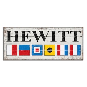 May include: A white wooden sign with the name "HEWITT" spelled out in black letters. Below the name are six nautical signal flags, each with a different color and pattern. The flags are red, white, blue, yellow, and black.