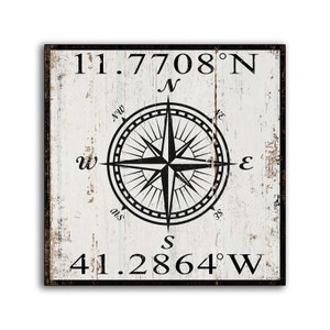 May include: A square, distressed wooden sign with a black border. The sign features a compass rose design in black, with the coordinates 11.7708°N and 41.2864°W in black text. The background is white with a weathered appearance.