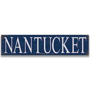 May include: A rectangular wooden sign with the word "NANTUCKET" in white capital letters against a distressed navy blue background. The sign has a rustic, weathered appearance, suggesting a coastal or nautical theme.