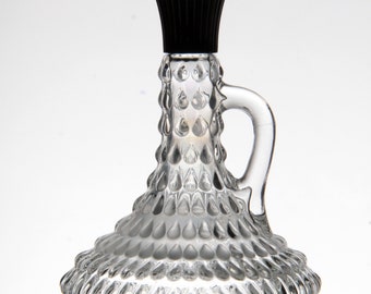 May include: A clear glass decorative pitcher with a black stopper. The pitcher has a handle and a textured, teardrop-shaped pattern. The design is repeated around the base and body of the pitcher.