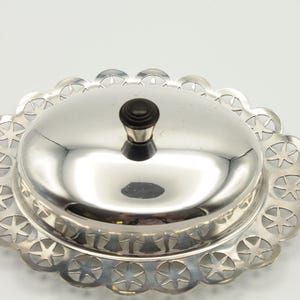 May include: A silver-colored butter dish with a domed lid and a decorative star-shaped pattern around the base. The lid has a black knob. The dish is oval-shaped and reflects the surrounding environment.