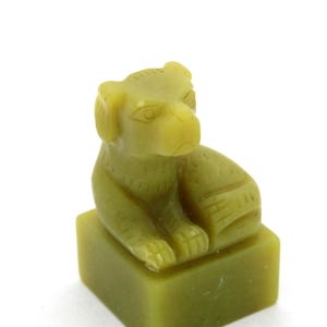 Puede incluir: Pequeña escultura de piedra tallada de color verde oliva que representa un perro. El perro está sentado sobre una base cuadrada. La escultura tiene detalles, incluyendo las orejas, ojos y patas del perro. Fondo blanco.