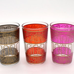 Puede incluir: Tres vasos de vidrio con diseños de patrones dorados. Los vasos son de vidrio transparente con un borde dorado y un diseño dorado. Los vasos están llenos de líquidos de diferentes colores: negro, naranja y rosa.
