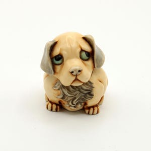 May include: A ceramic puppy figurine with a mournful expression. The puppy is beige with grey ears and green eyes. It has a detailed grey and beige fur. The puppy is in a sitting position.