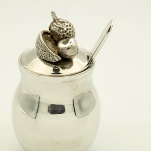 May include: A silver sugar bowl with an acorn-shaped lid and a small spoon. The bowl has two decorative shapes on the front. The bowl is made of a shiny, reflective metal.