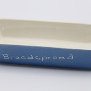 May include: A rectangular breadspread dish with a blue exterior and a cream-colored interior. The word "Breadspread" is written in white script on the blue side. The dish is made of ceramic.