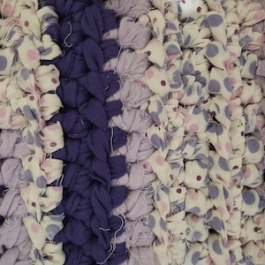May include: Close-up of a handmade rug with a braided design. The rug features a mix of fabrics in various shades of purple, cream, and white. Some sections have a polka dot pattern in pink and purple.