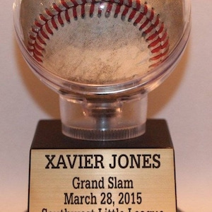 May include: A baseball encased in a clear plastic dome sits on a black base with a gold plaque. The plaque reads "XAVIER JONES Grand Slam March 28, 2015 Southwest Little League The Cyclones".
