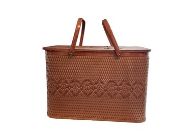 May include: A brown woven picnic basket with a wooden handle and lid. The basket has a rectangular shape with a decorative woven pattern on the front. The handle is arched, and the lid is secured with a latch.